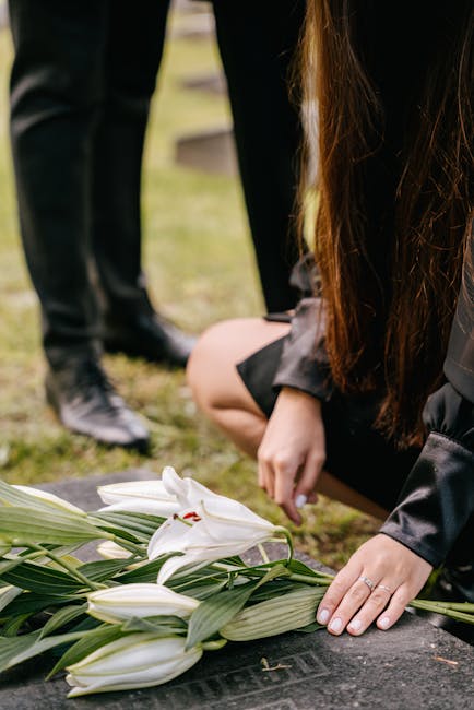 A person with long brown hair, dressed in dark clothing, is kneeling on the grass at a funeral setting, carefully arranging white lilies with green leaves on a dark stone memorial. The arrangement inc