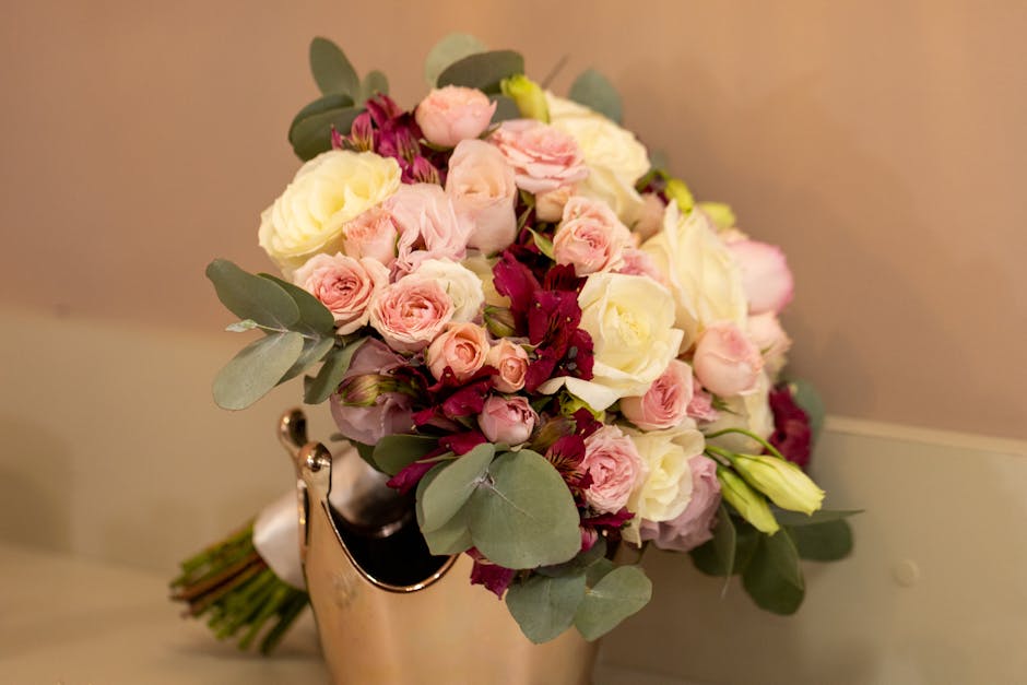 A close-up of a lush floral bouquet arranged with pale pink roses, white ranunculus, and deep burgundy flowers, accented with eucalyptus leaves. The bouquet is wrapped in cream-colored wrapping paper 