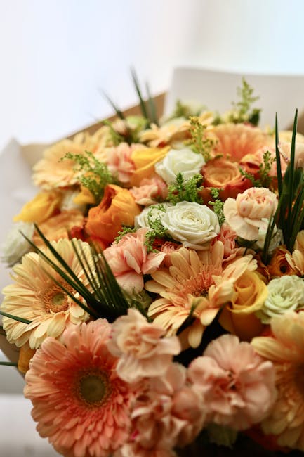 A vibrant bouquet featuring a mix of fresh flowers including pale peach gerbera daisies, soft pink ranunculus, creamy white roses, and peach-orange lisianthus, accented with green foliage and grass-li