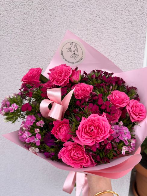 A vibrant floral bouquet consisting of large, bright pink roses with fully opened petals, complemented by smaller purple and pink blossoms, arranged in a lush, dense display. The flowers are wrapped i