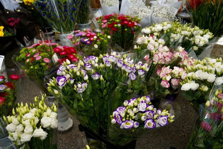 A vibrant display of fresh flowers arranged in bouquets, featuring purple and white eustoma with ruffled petals, complemented by pink and white spray roses, all wrapped in clear plastic. The flowers a