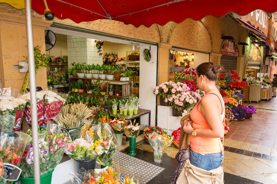 A woman stands outside a flower shop near Regents Park, browsing a vibrant display of fresh flowers and floral arrangements. The bouquet she is holding features pink roses, red carnations, and purple