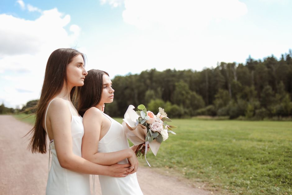 Two young women with long brown hair, dressed in white sleeveless dresses, stand outdoors on a gravel path with a lush green field and trees in the background under a partly cloudy sky. The woman on t