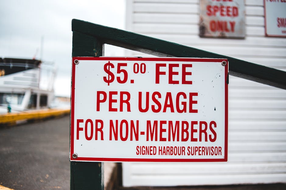 Close-up of a white rectangular sign with red text that reads '$5.00 FEE PER USAGE FOR NON-MEMBERS' and is signed by the Harbour Supervisor, mounted on a dark green metal post. In the background, ther